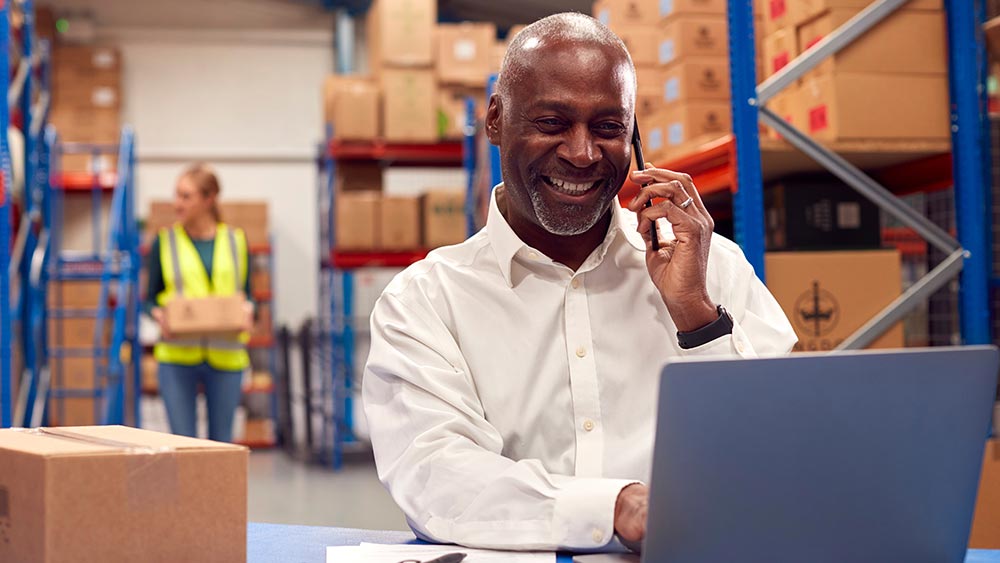 Amtivo (Formerly Certification Europe and EQA) - ISO Certification Transfer - A person in a warehouse on a laptop while taking a business call.